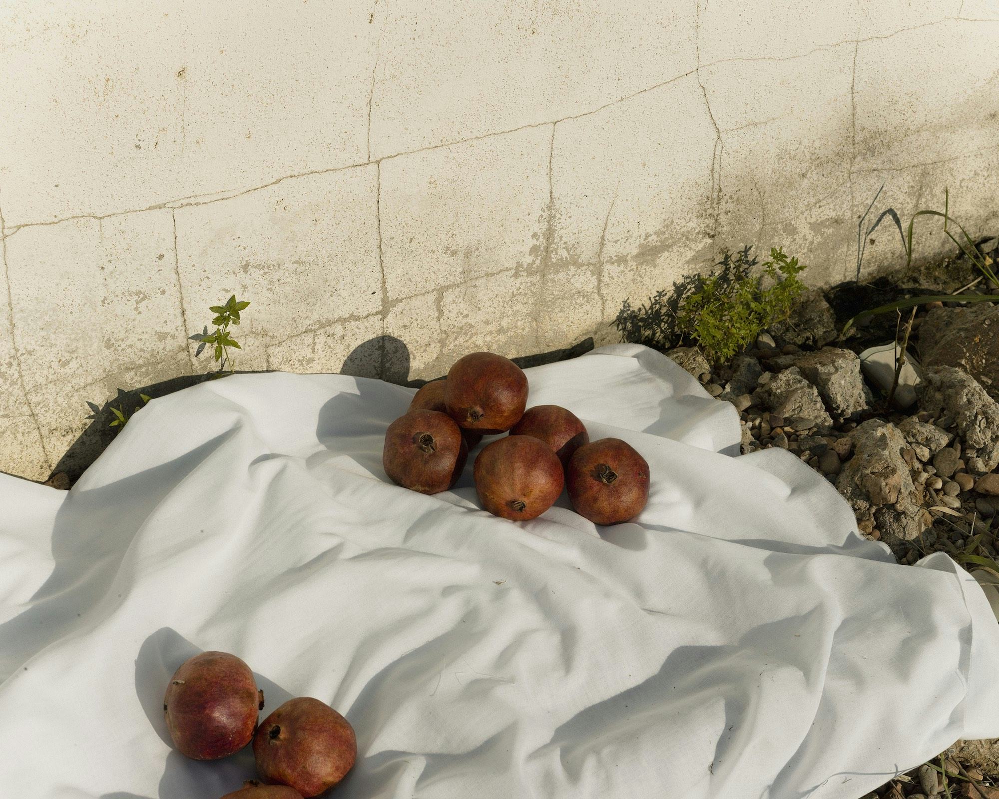 Image of pomegranates on a white table cloth, on a rocky surface in front of a beige, cracked wall. © Felipe Romero Beltrán