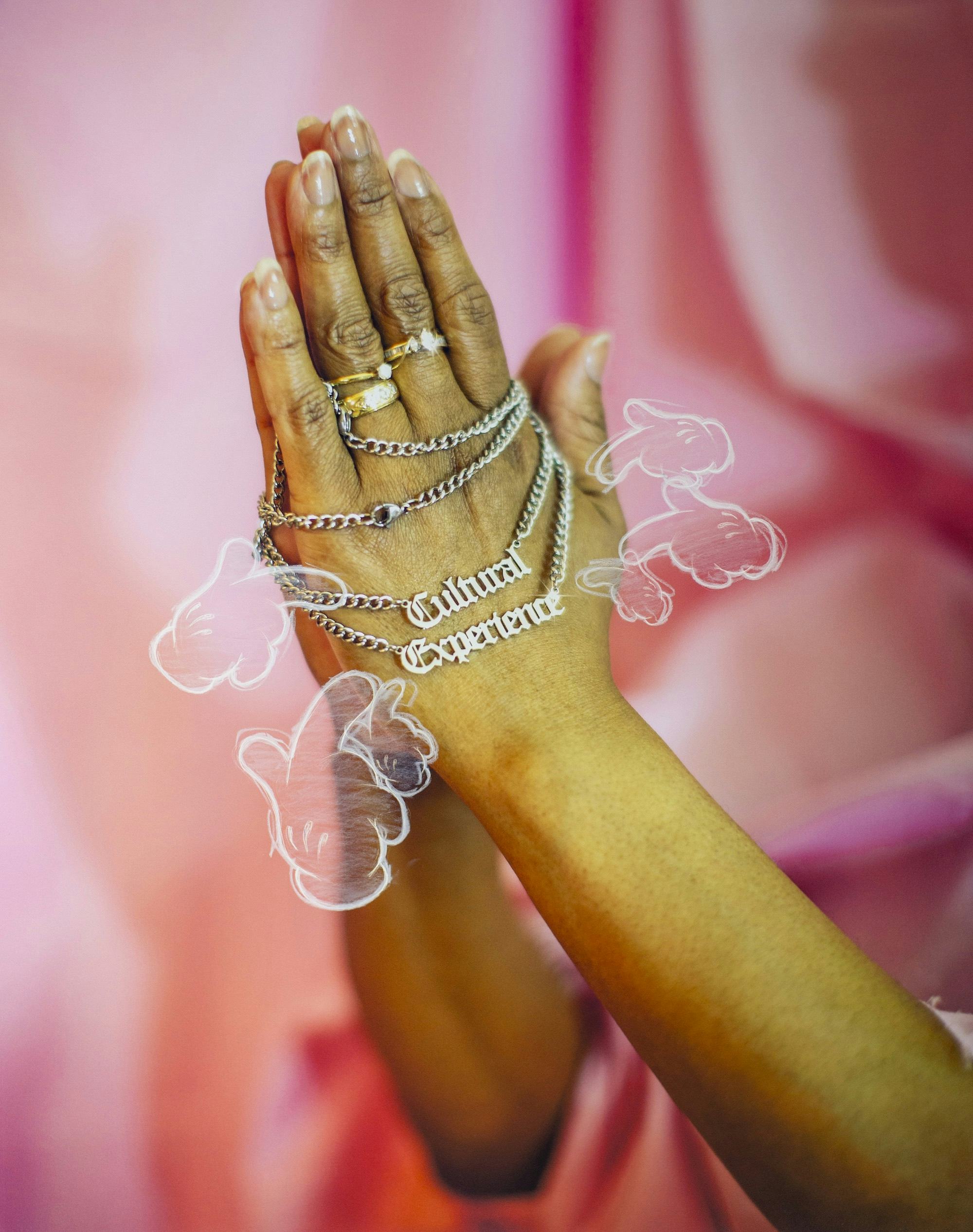 Photo of a pair of hands with rings in prayer gesture, holding a chain that spells 'Cultural Experience', in front of a pink silky background. © André Ramos-Woodard