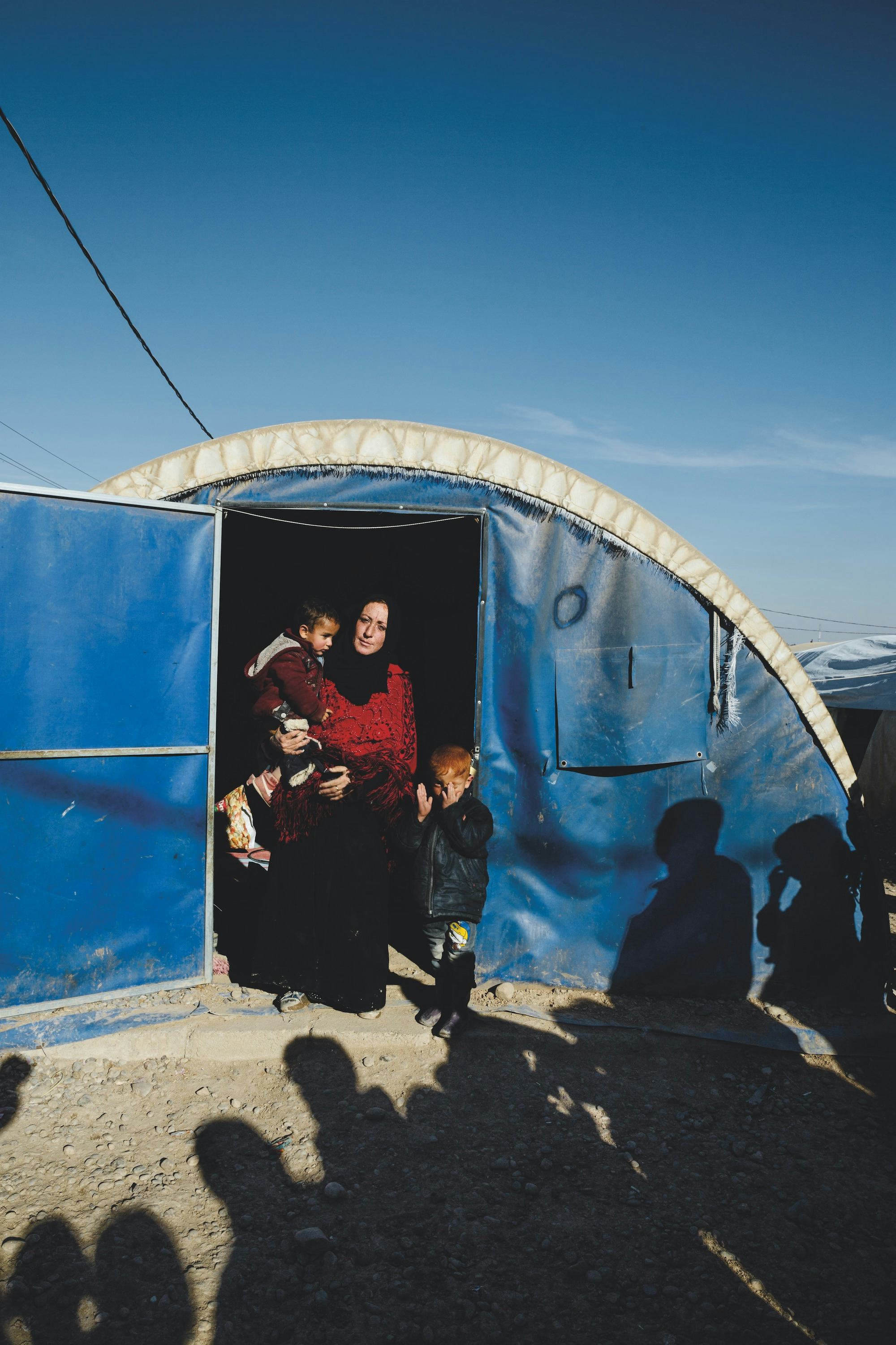A woman carrying a child and one child at her feet, in the door opening of a refugee / shelter tent.