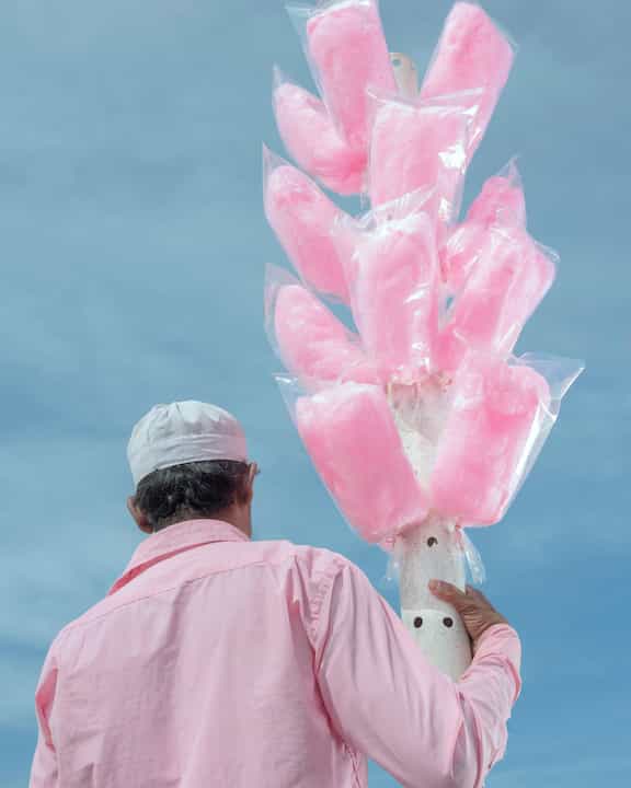 A person in a pink shirt and cap holds a pole with multiple bags of pink cotton candy against a blue sky.