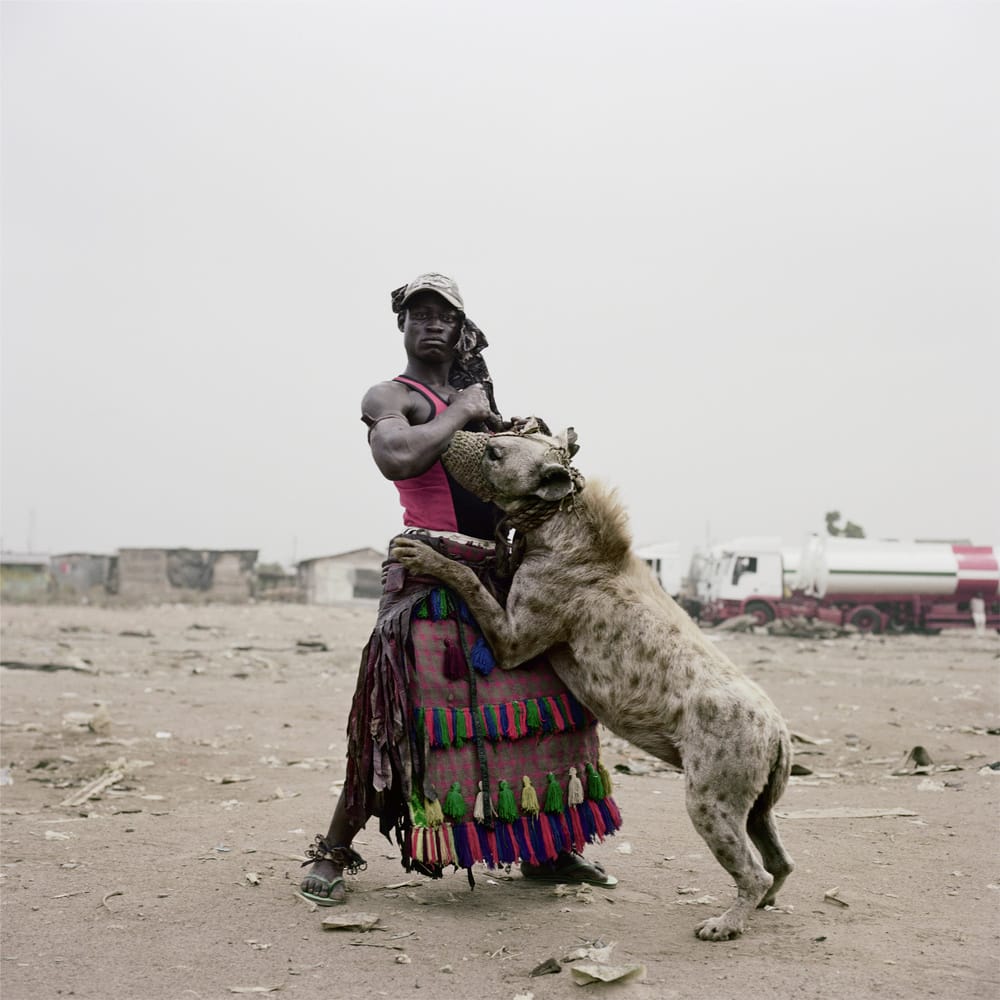 Nigeriaan staat in een verlaten landschap met op de achtergrond wat hutjes en een tankauto. Een hyena staat tegen hem aangeleund.