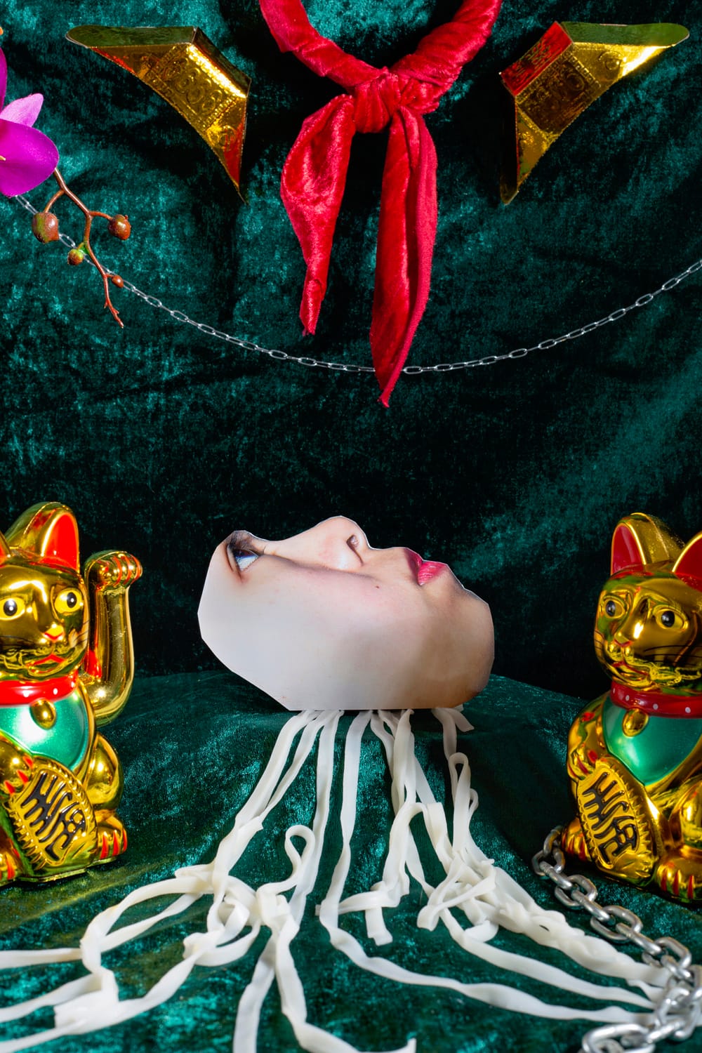 A surreal scene with a mask-like face on noodles, surrounded by two golden cat figurines, red fabric, and decorative elements on a green background.