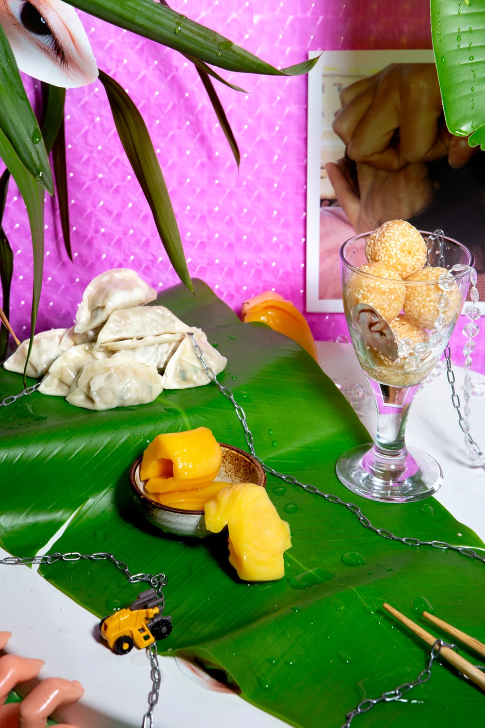 A vibrant still life with dumplings, mango slices, sesame balls in a glass, toy truck, and chains on banana leaves against a pink background.