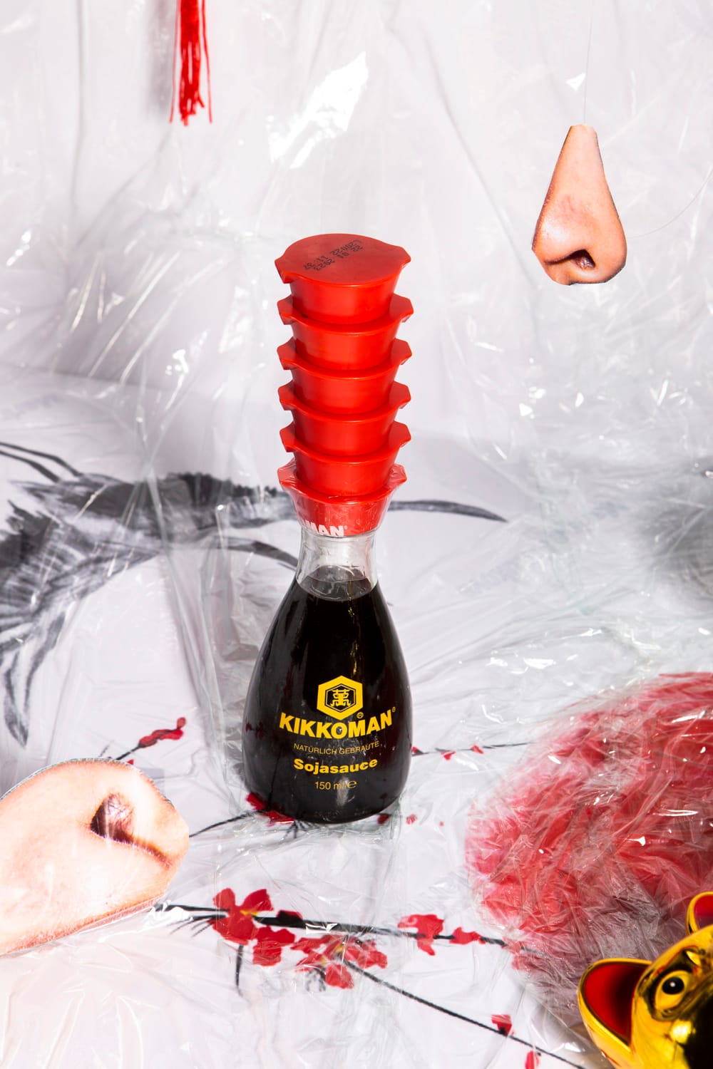 A Kikkoman soy sauce bottle with a stack of red caps, surrounded by abstract art featuring noses and red floral elements.
