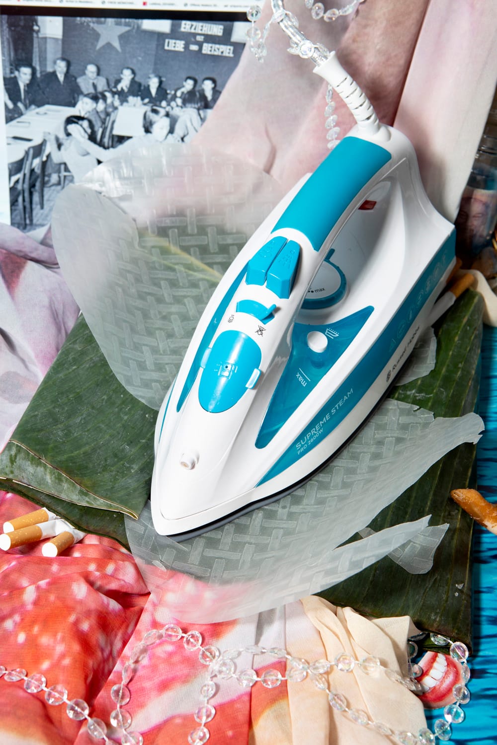 A blue and white steam iron on a textured rice paper surface, surrounded by various fabrics, pills, and a vintage photo in the background.