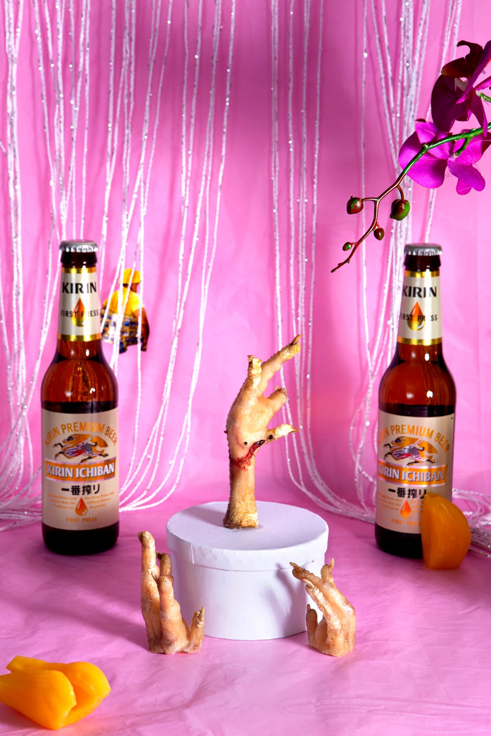 Two Kirin Ichiban beer bottles flank a chicken foot on a pedestal, with pink backdrop, orchids, and orange petals scattered around.