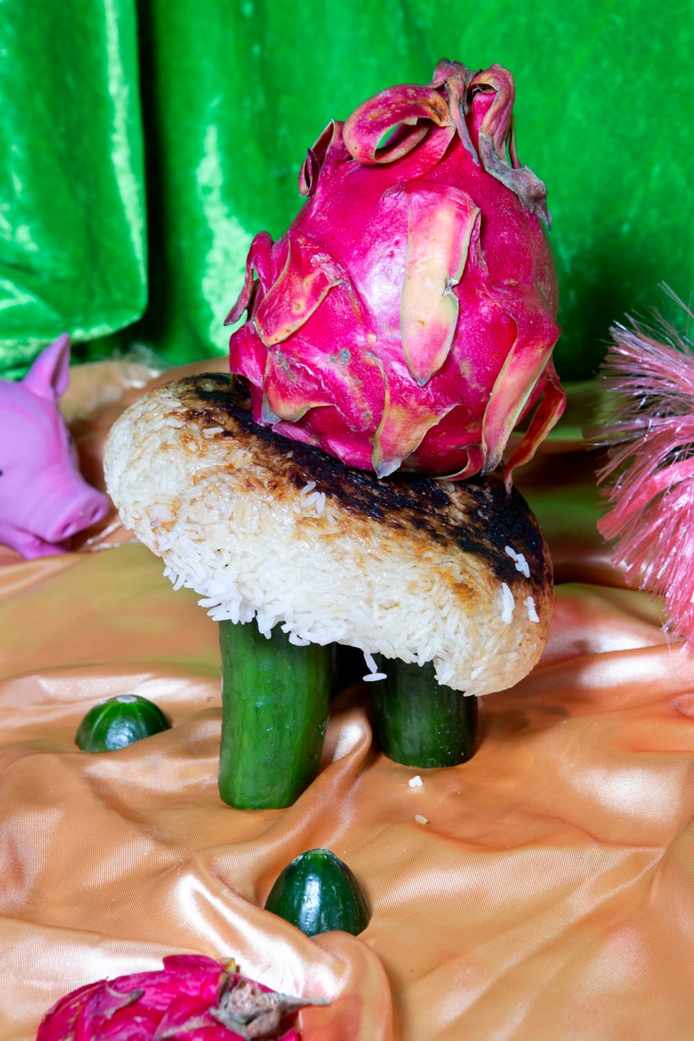 A dragon fruit on a toasted rice patty with cucumber legs, surrounded by small cucumbers and a pink plastic pig on a silky orange fabric.