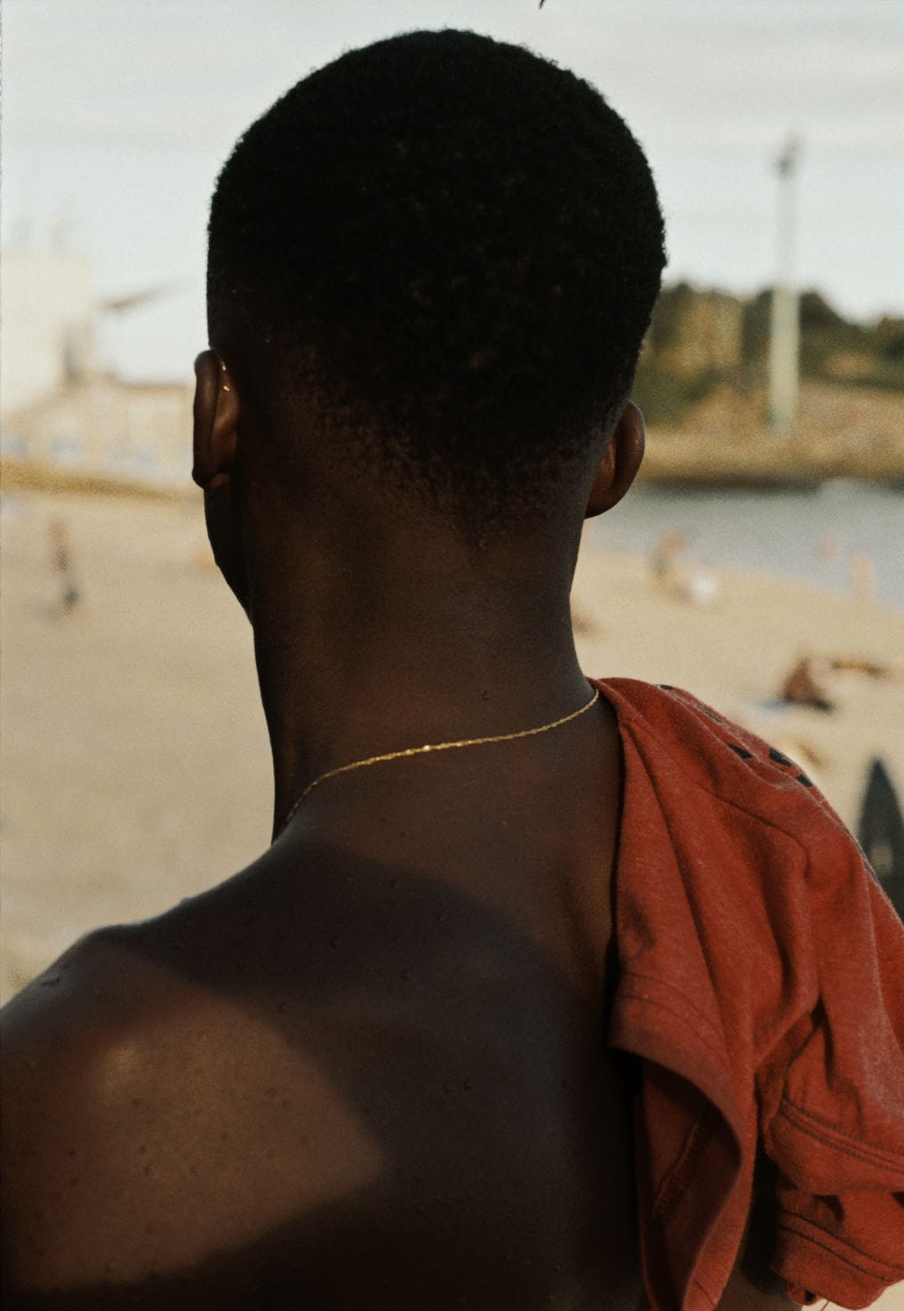 A person with short hair stands on a beach, facing away, with a red shirt draped over their shoulder and a thin necklace visible.