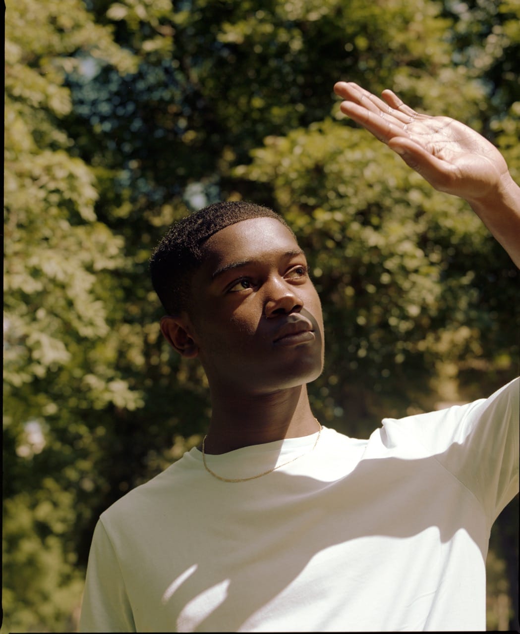 A person in a white shirt shields their eyes from the sun with one hand, standing outdoors with trees in the background.