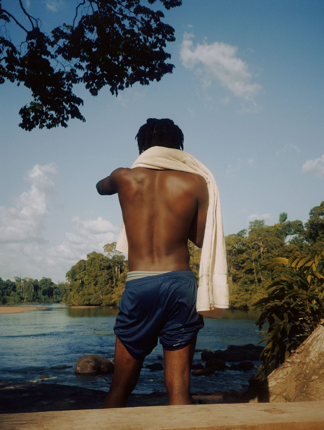Person with a towel over their shoulder stands by a river, surrounded by lush greenery and a clear blue sky.