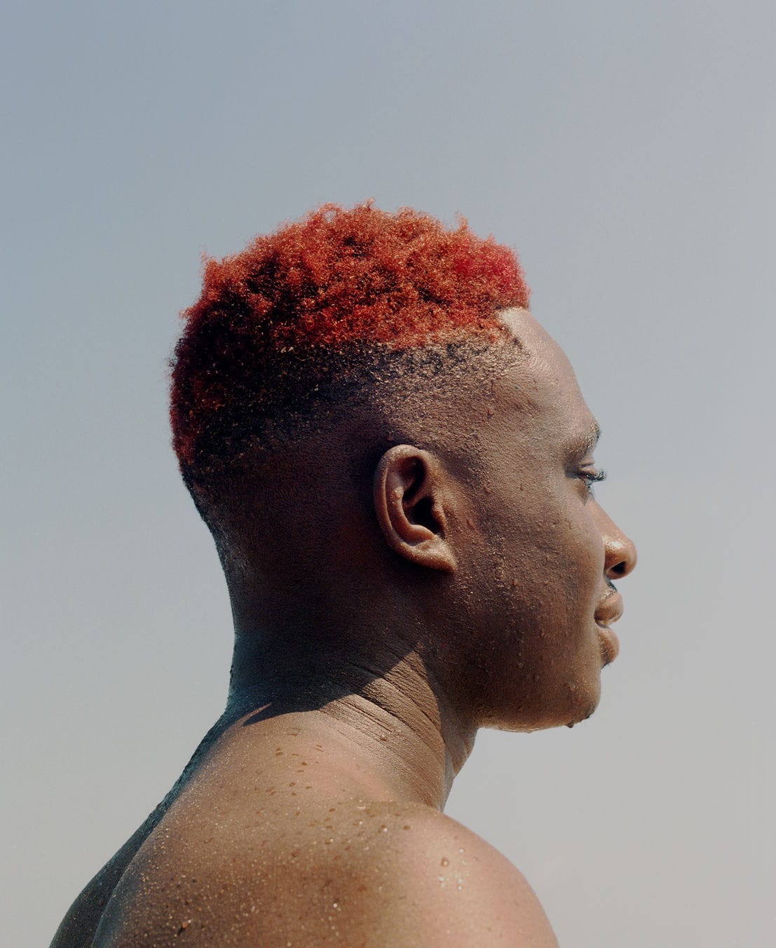 Profile of a person with short, bright red hair, looking to the right against a clear blue sky. Skin glistens as if wet.