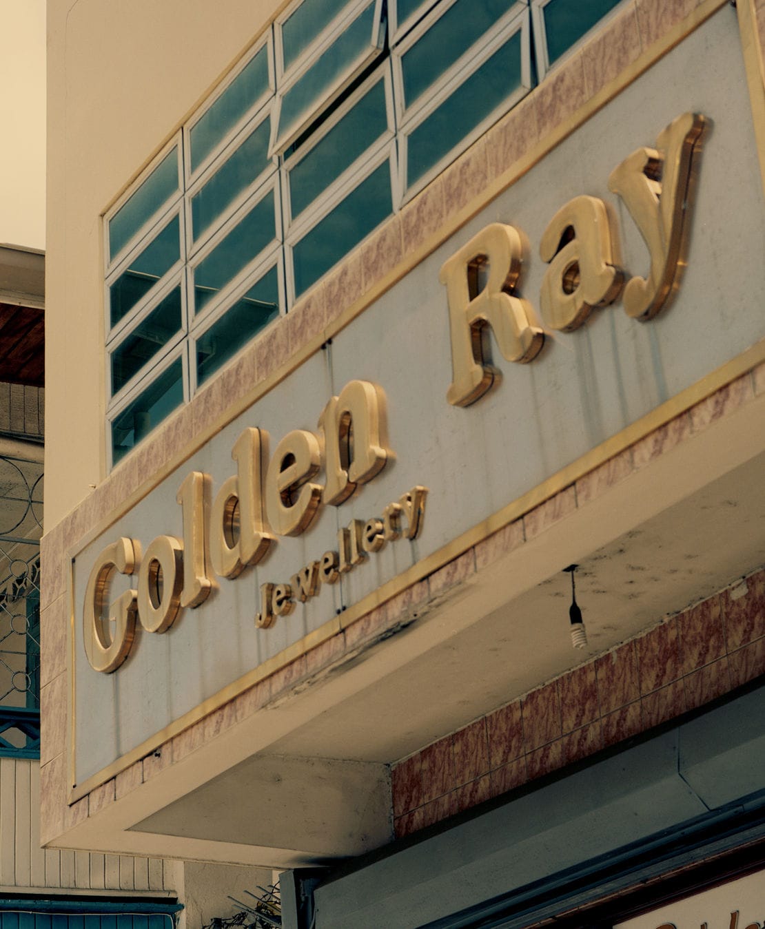 Sign for "Golden Ray Jewellery" with gold lettering on a building facade, featuring large windows above.