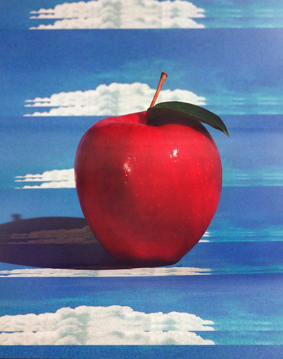 A shiny red apple with a green leaf sits against a blue sky backdrop with white clouds, casting a shadow on the surface.