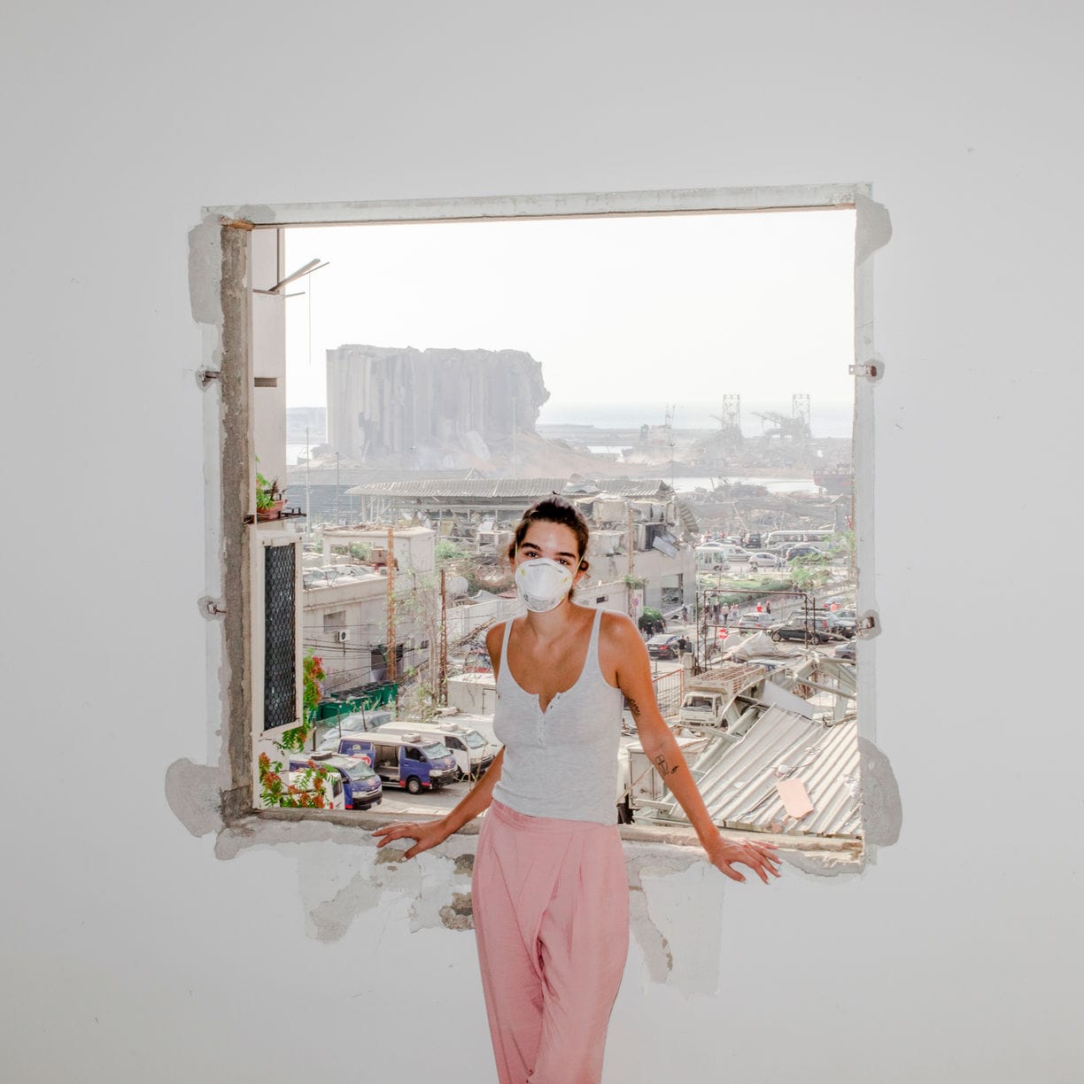 Woman in a white mask and pink pants stands in a building's window frame overlooking a cityscape with damaged structures.
