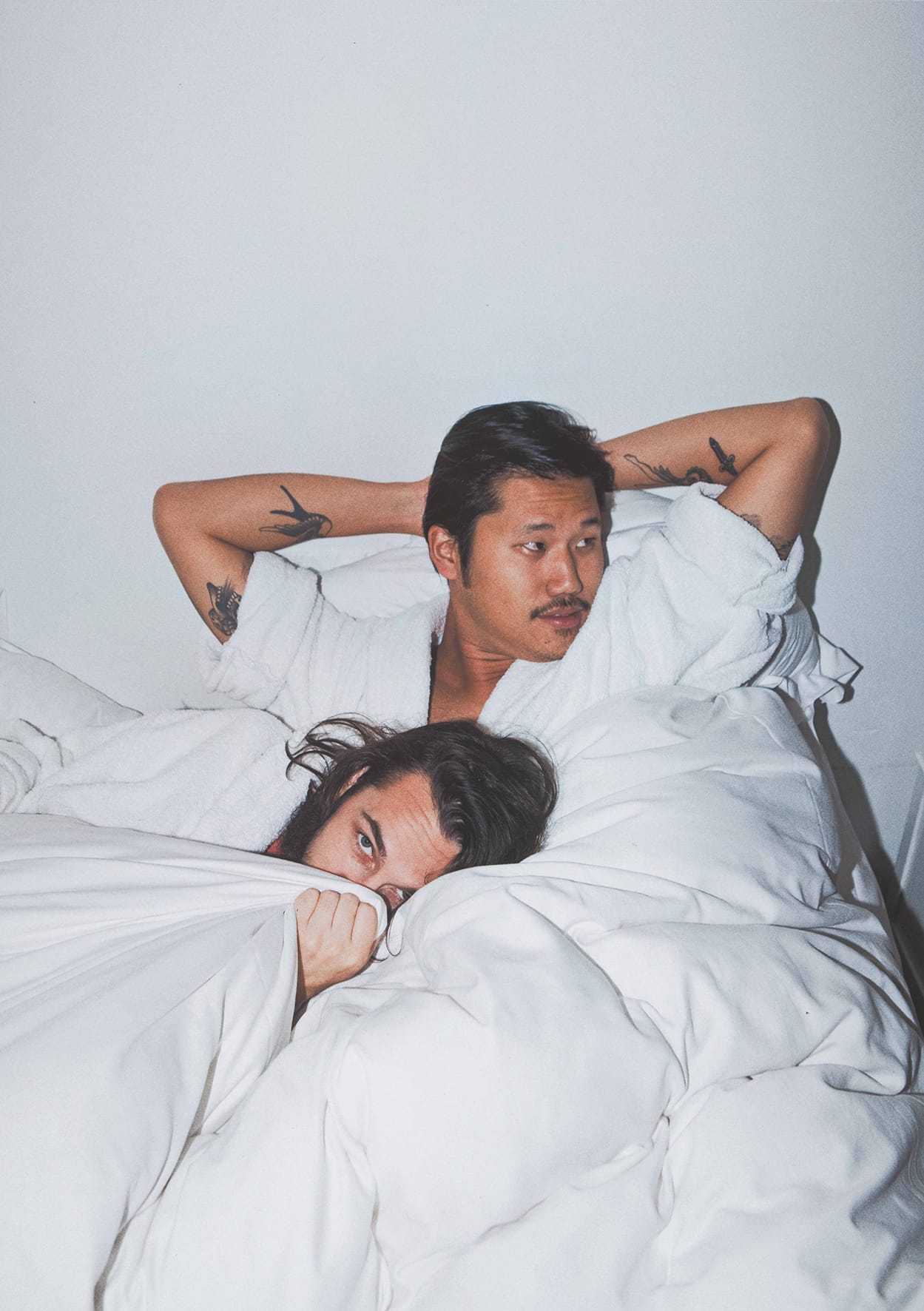A portrait of two musicians relaxing in bed, one looking away and the other looking into the camera from behind the sheets.