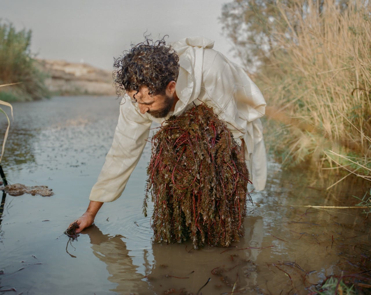 A person in a white coat and seaweed skirt bends down to touch water in a marshy area, surrounded by reeds and soft lighting.