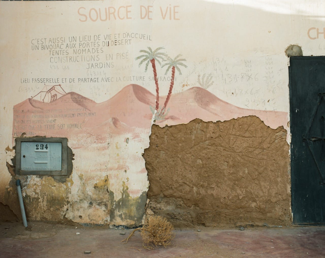 Wall with mural of desert scene and palm trees, faded French writing, cracked plaster, mailbox, and a tumbleweed on the ground.
