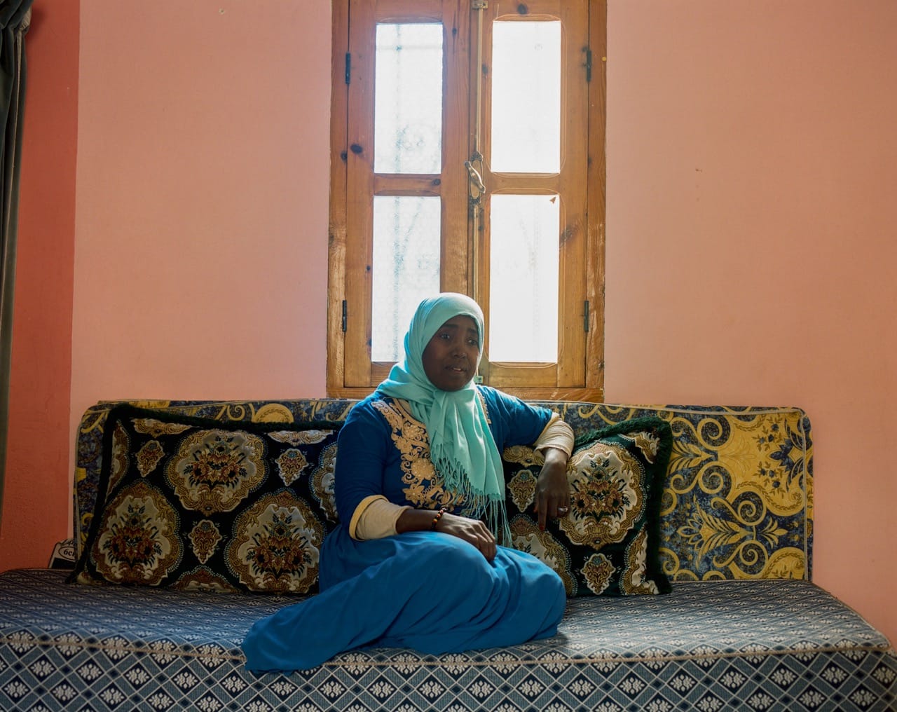 A woman in a blue dress and headscarf sits on a patterned couch in a room with pink walls and a wooden window.