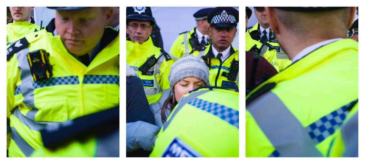 Police officers in high-visibility jackets interact with a protester wearing a gray beanie. The scene appears tense and crowded.