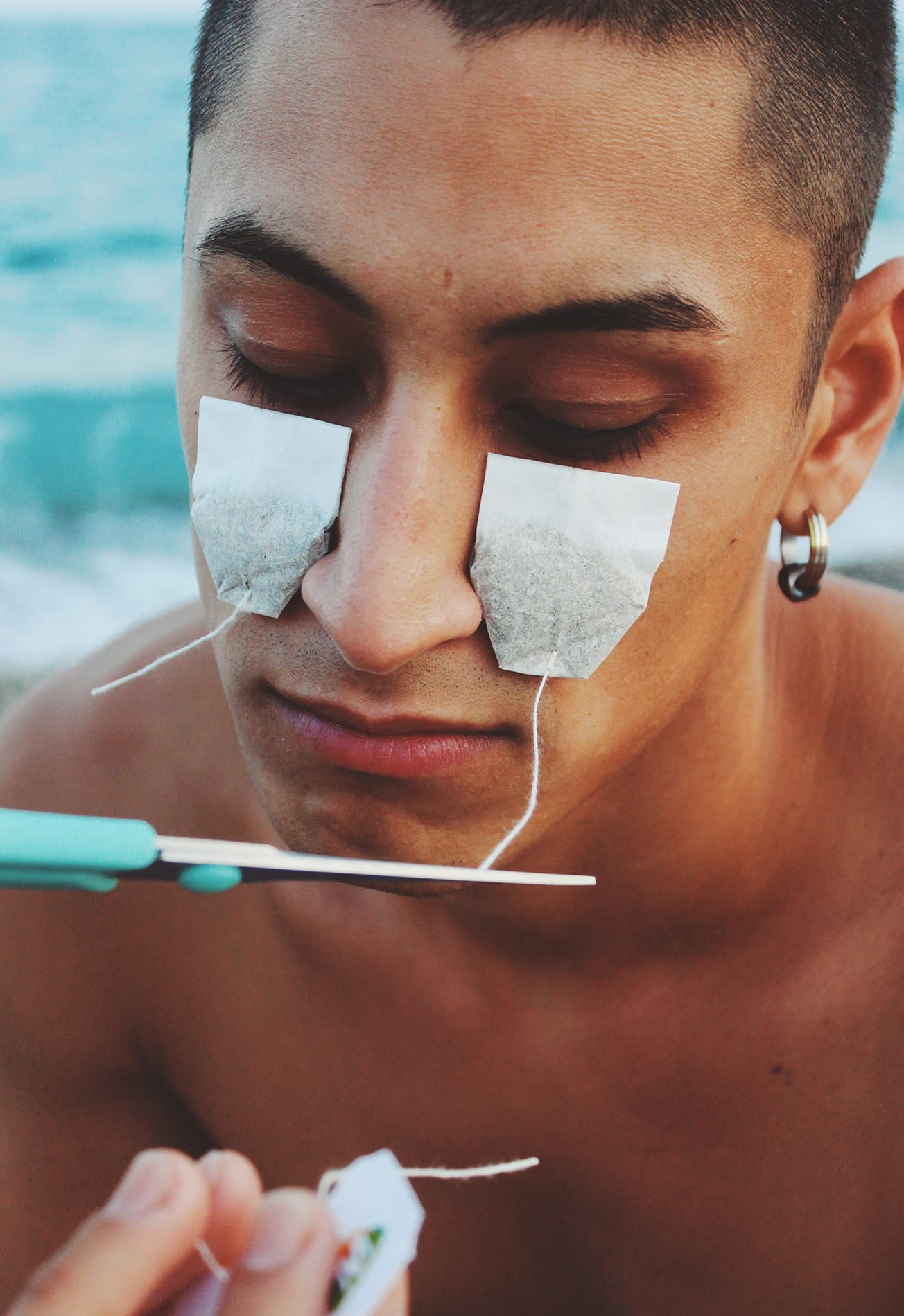 Portrait of a man with tea bags under his eyes, a pair of scissors is seen cutting of the tags. © Alp Peker