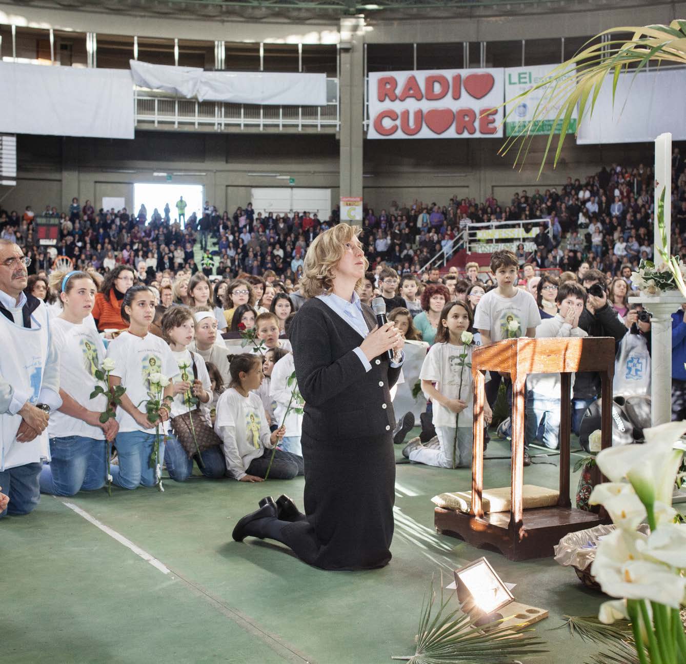 A woman kneels in prayer on a gym floor surrounded by people, some holding flowers, in a large indoor gathering.