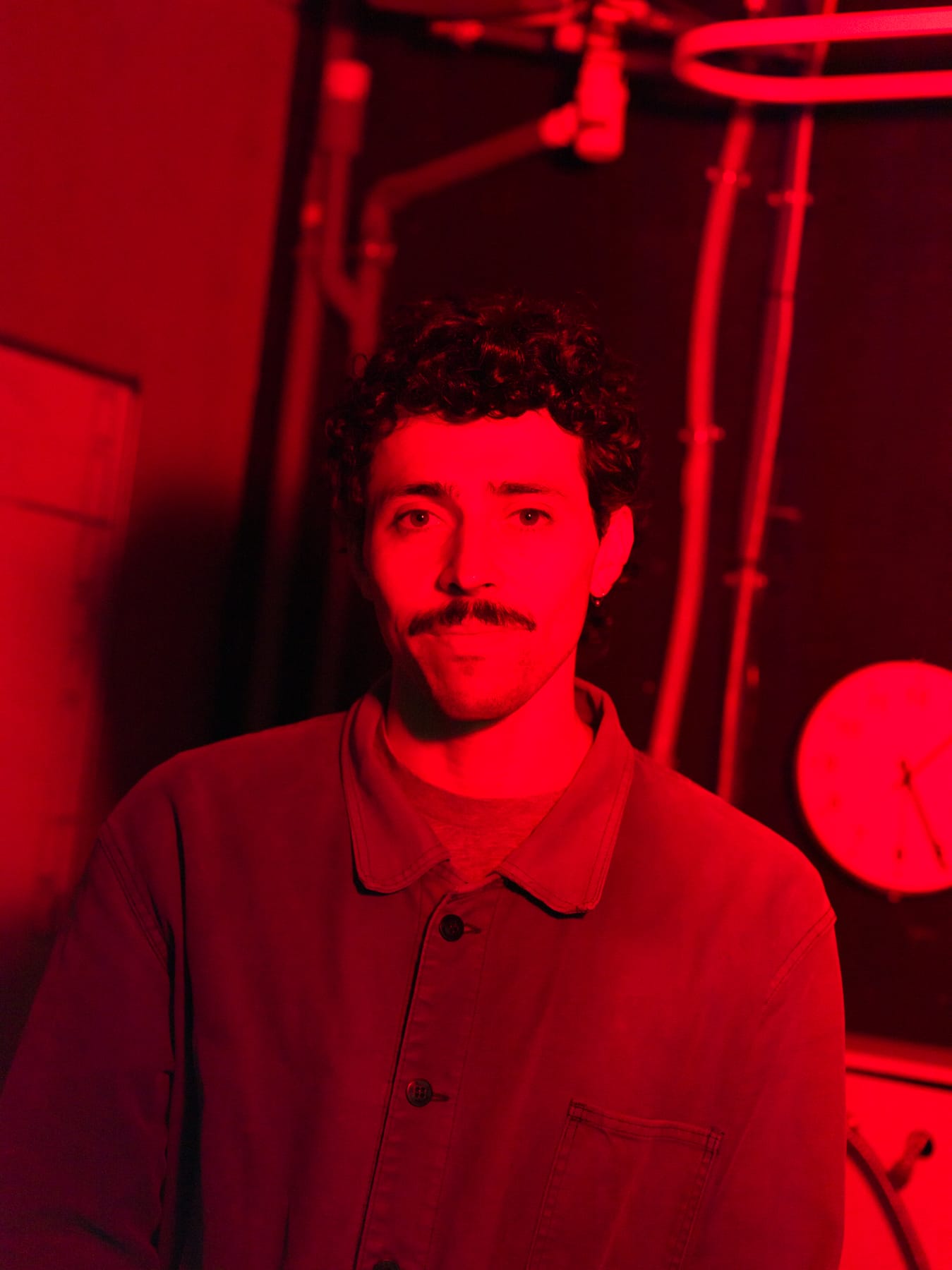 A person with curly hair and a mustache stands in a red-lit room with industrial pipes and a clock in the background.