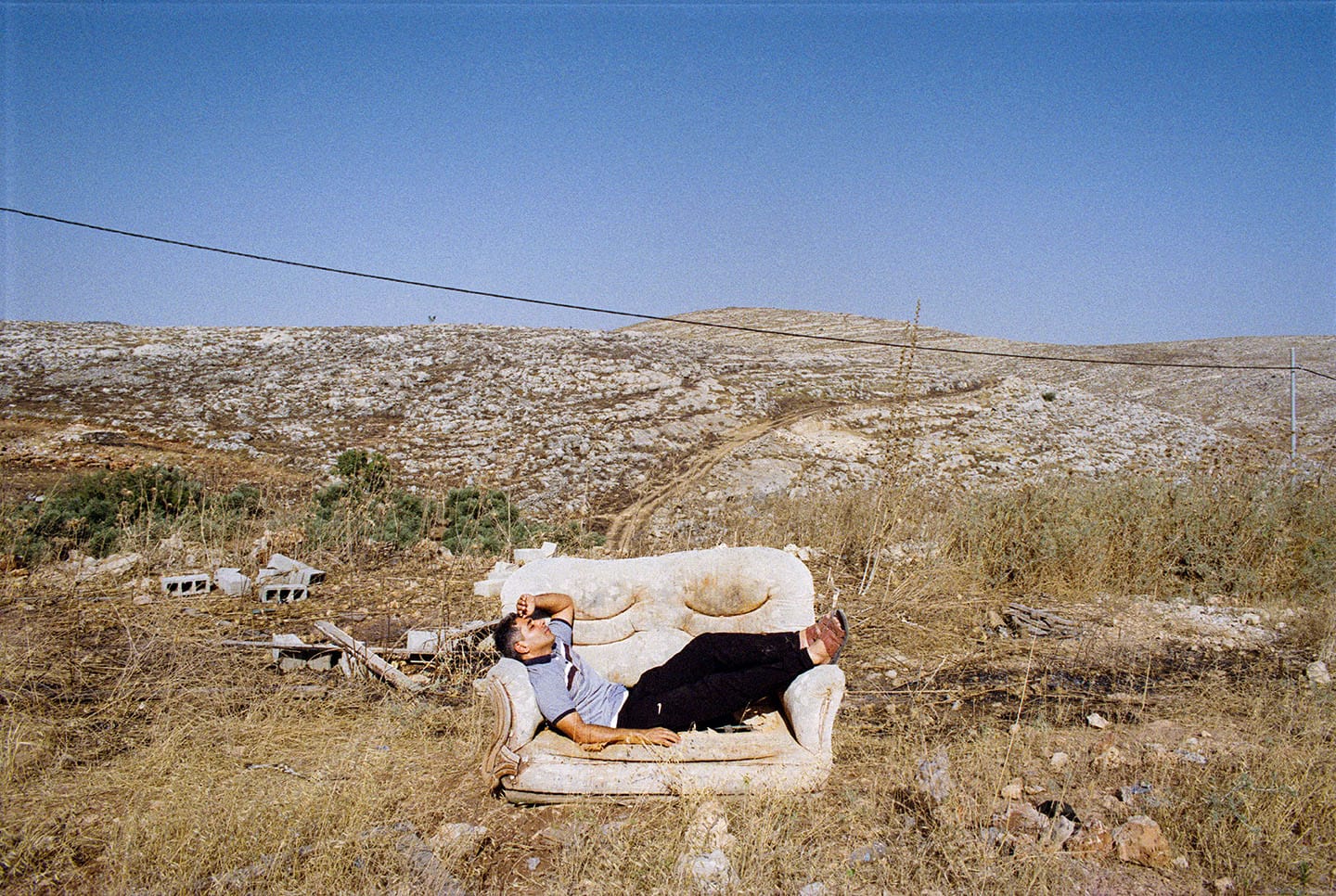 Person lounging on a worn-out sofa in a dry, rocky landscape under a clear blue sky, with scattered shrubs and distant hills.