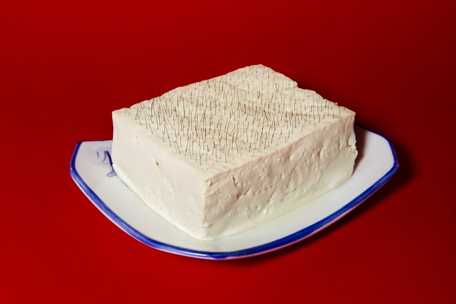 A white tofu block with numerous small pins on top, placed on a decorative plate against a solid red background.