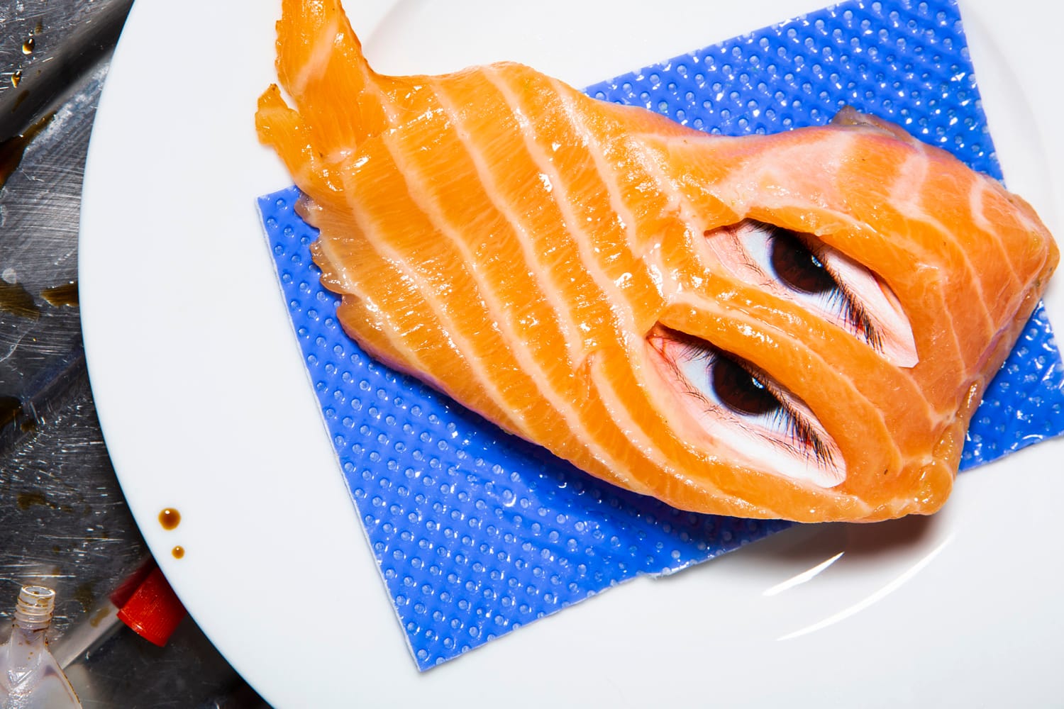 Salmon fillet with human eyes on a white plate, placed on a blue textured mat. Surreal and artistic composition.