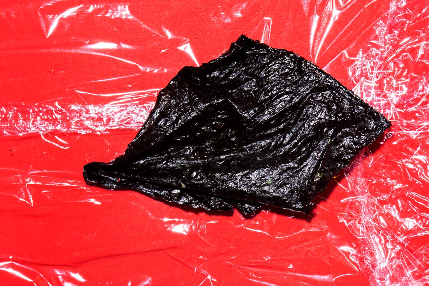 A piece of dark seaweed on clear plastic wrap against a bright red background.