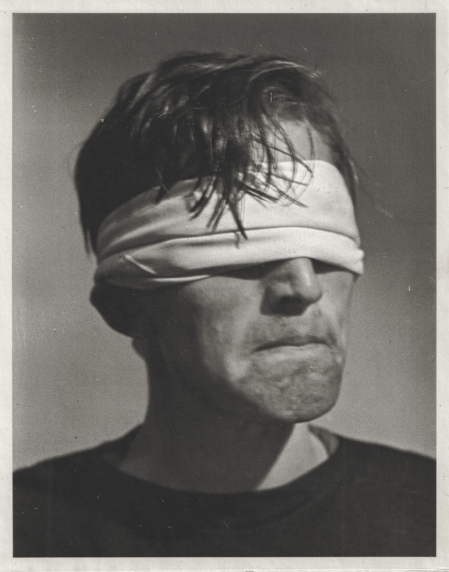 A person with tousled hair and a bandage covering their eyes, appearing contemplative against a plain background.