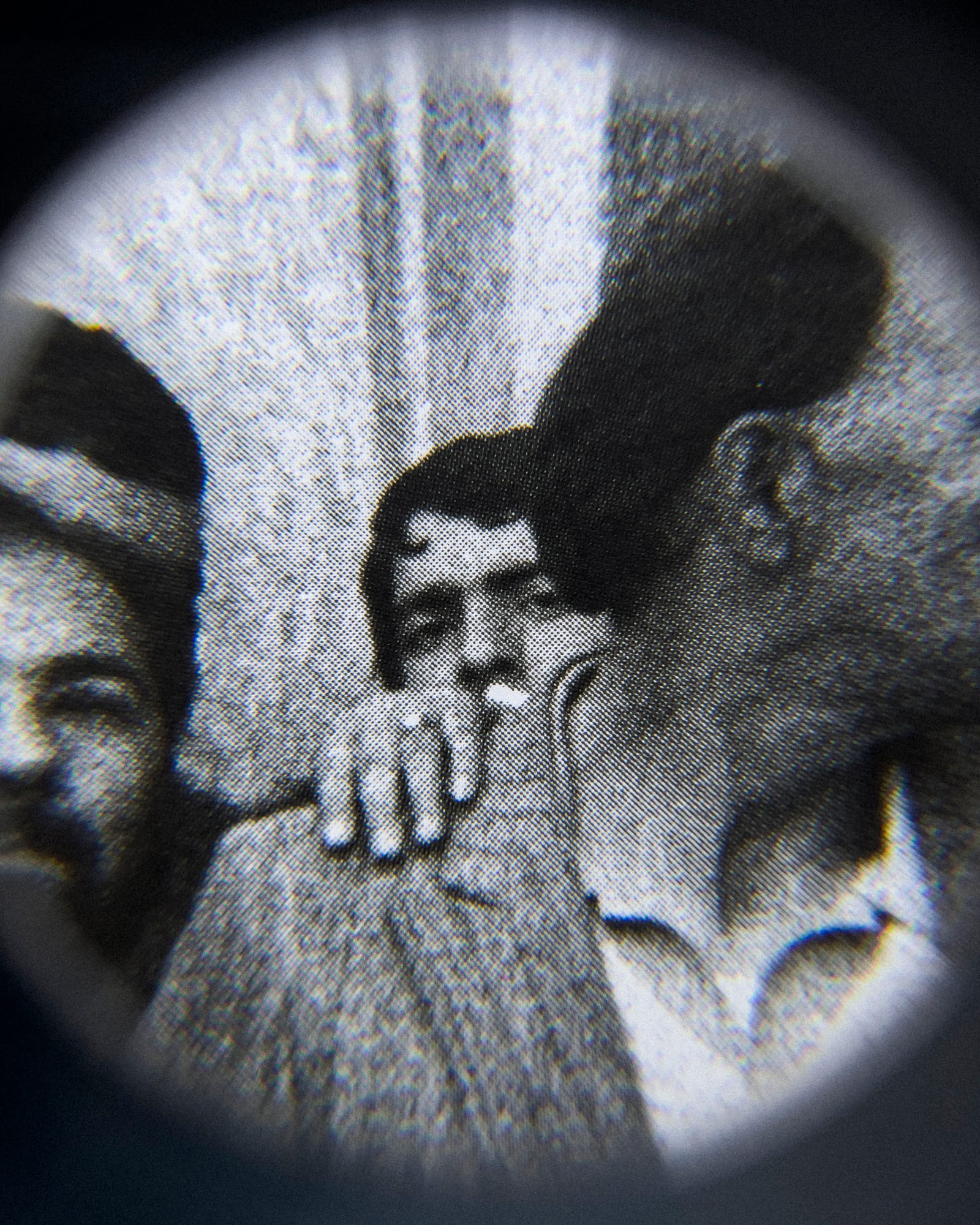 Black and white close-up image of an existing photograph, showing a man peeking into the camera from behind another man. Shot through a magnifying lens © Amin Yousefi