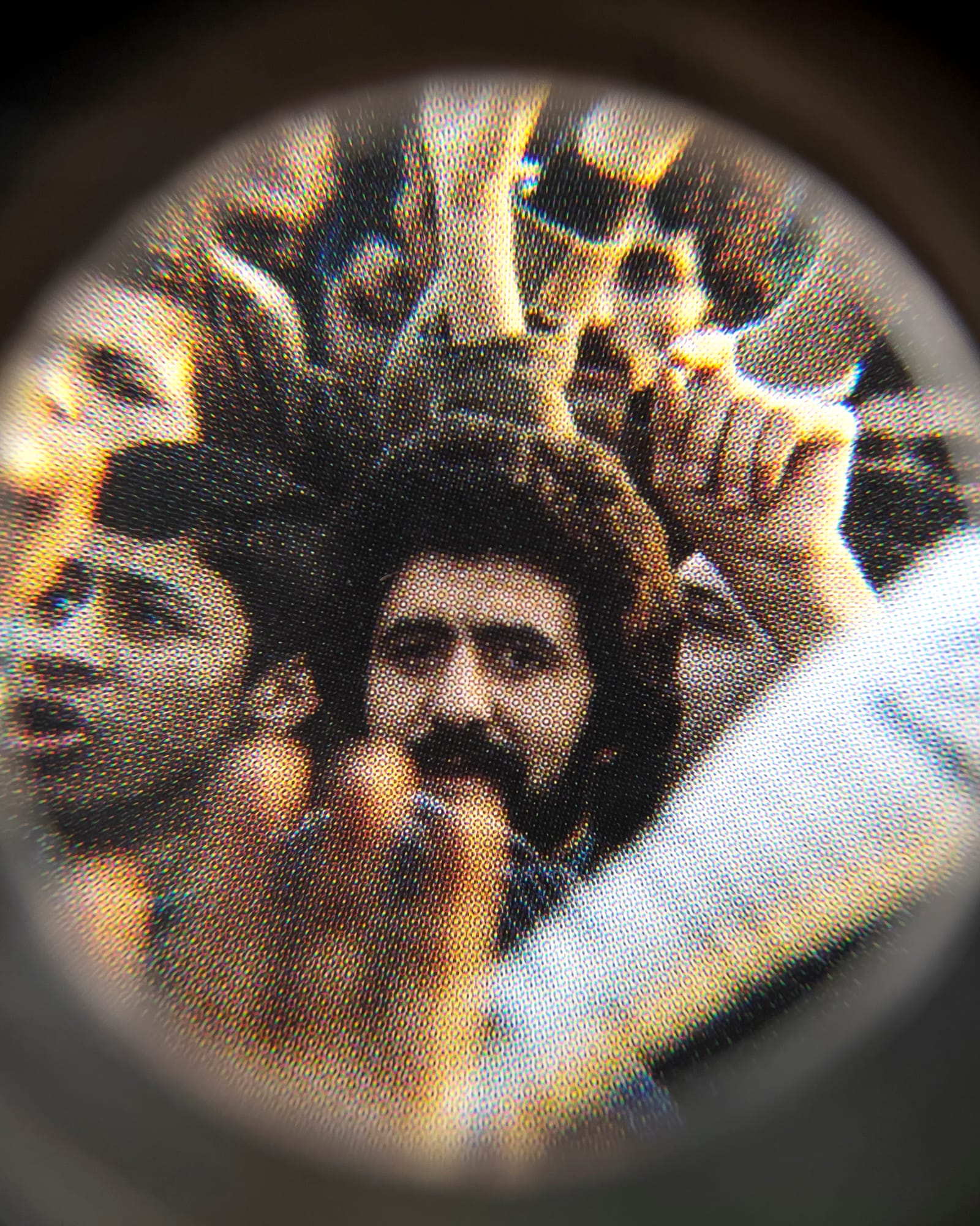 Coloured close-up image of an existing photograph, showing a man in the middle of a protesting crowd staring into the camera. Shot through a magnifying lens © Amin Yousefi
