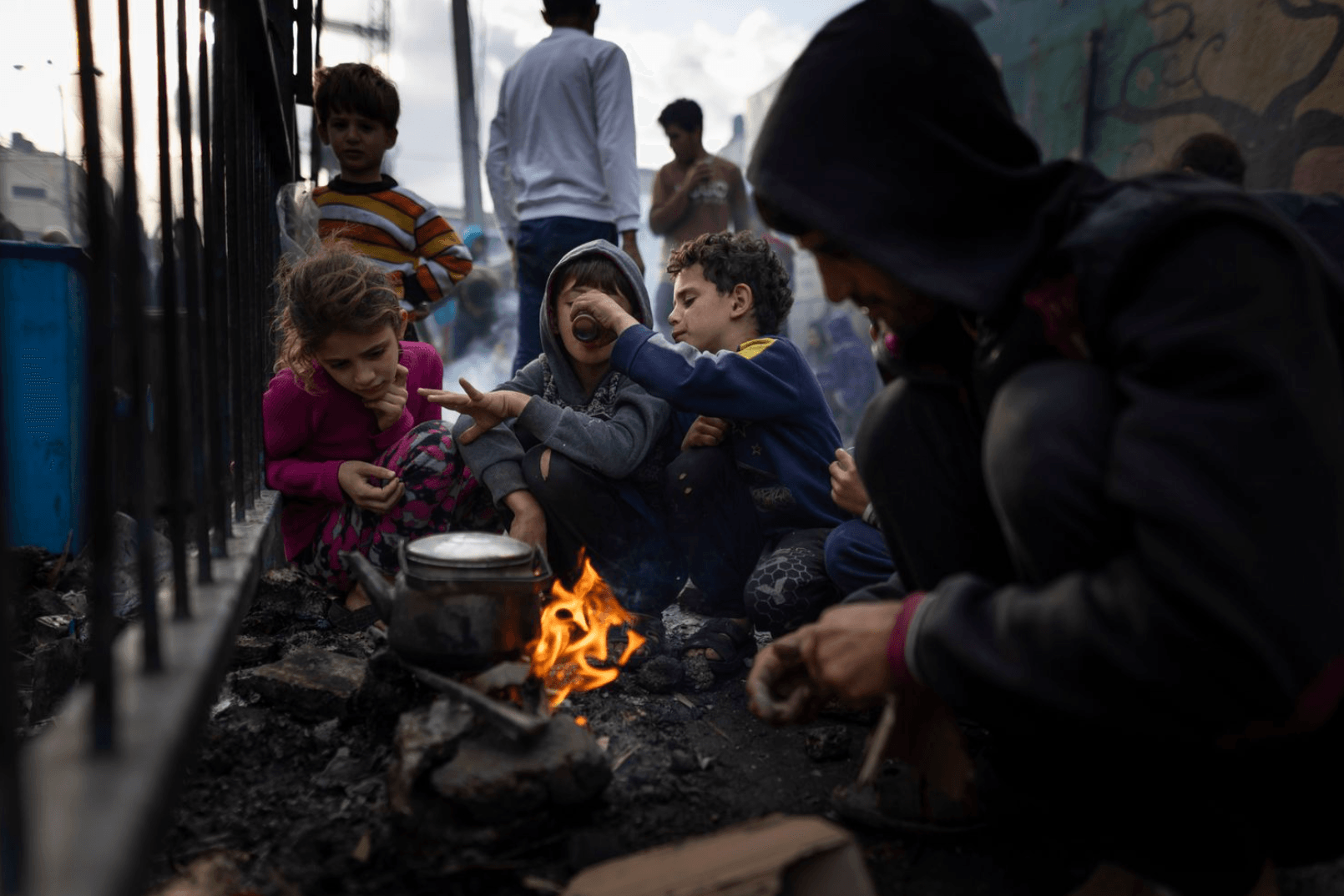 Children and a man sit around a small outdoor fire, boiling water in a pot. One child drinks from a cup while others watch.
