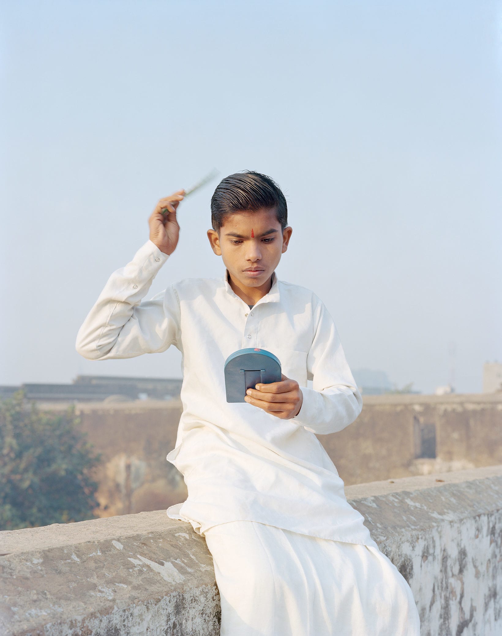 Beeld van Indiase jongen in witte kleding op een muurtje terwijl hij zijn haren kamt.