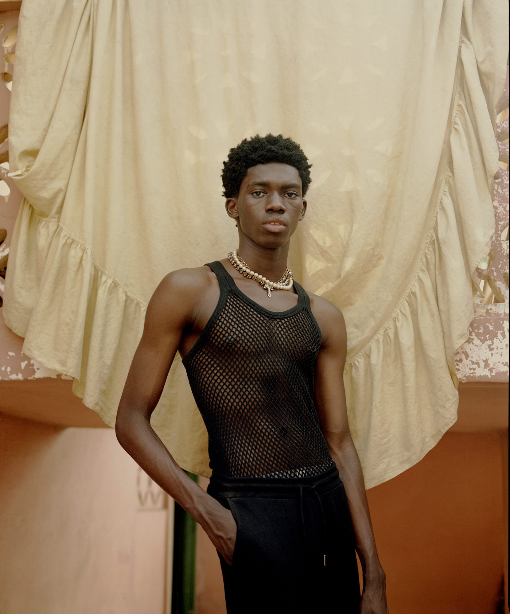 Young man in a black mesh tank top and black pants stands confidently, with a beige draped fabric as a backdrop.