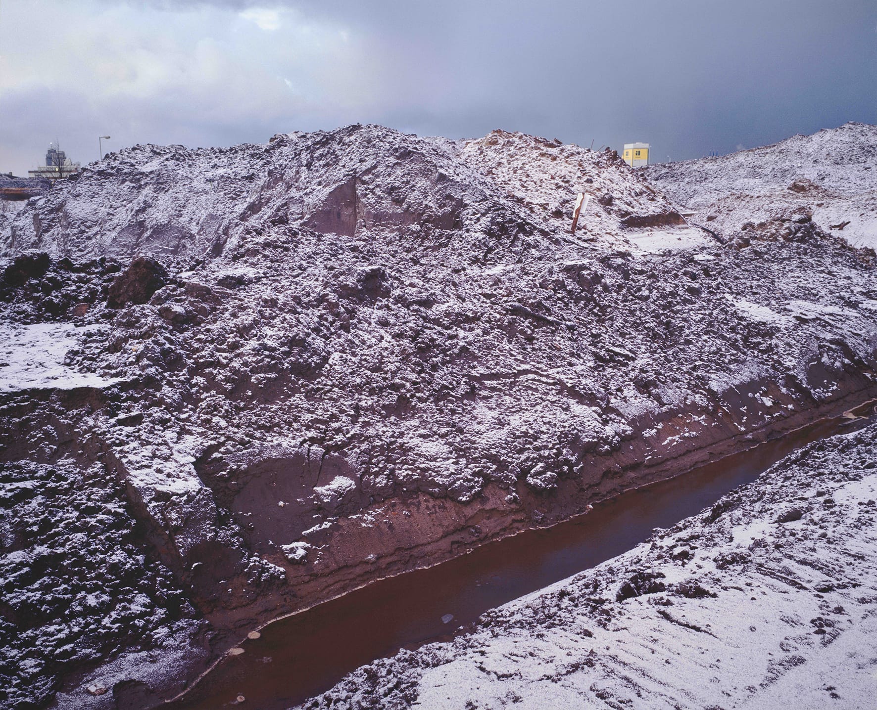 Besneeuwde zandhoop met op achtergrond twee bouwkeetjes, de afbeelding lijkt op een berglandschap