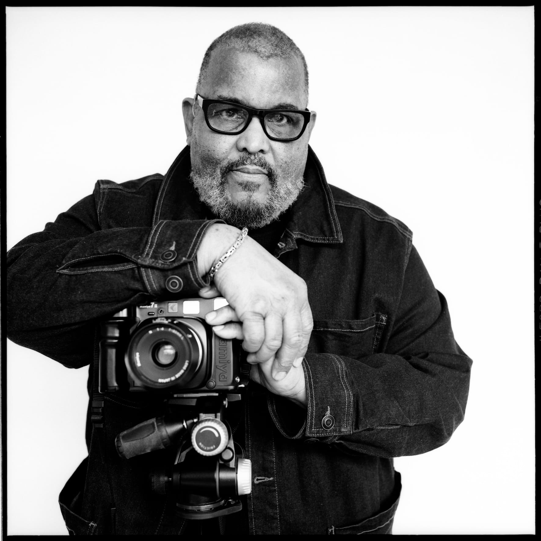 A person in a denim jacket holds a camera on a tripod, against a plain background, with a confident expression. Black and white photograph.