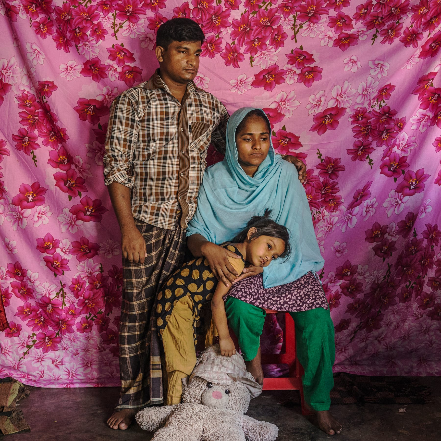 A family of three poses in front of a pink floral backdrop; the mother sits with a child resting on her lap, and the father stands beside them.