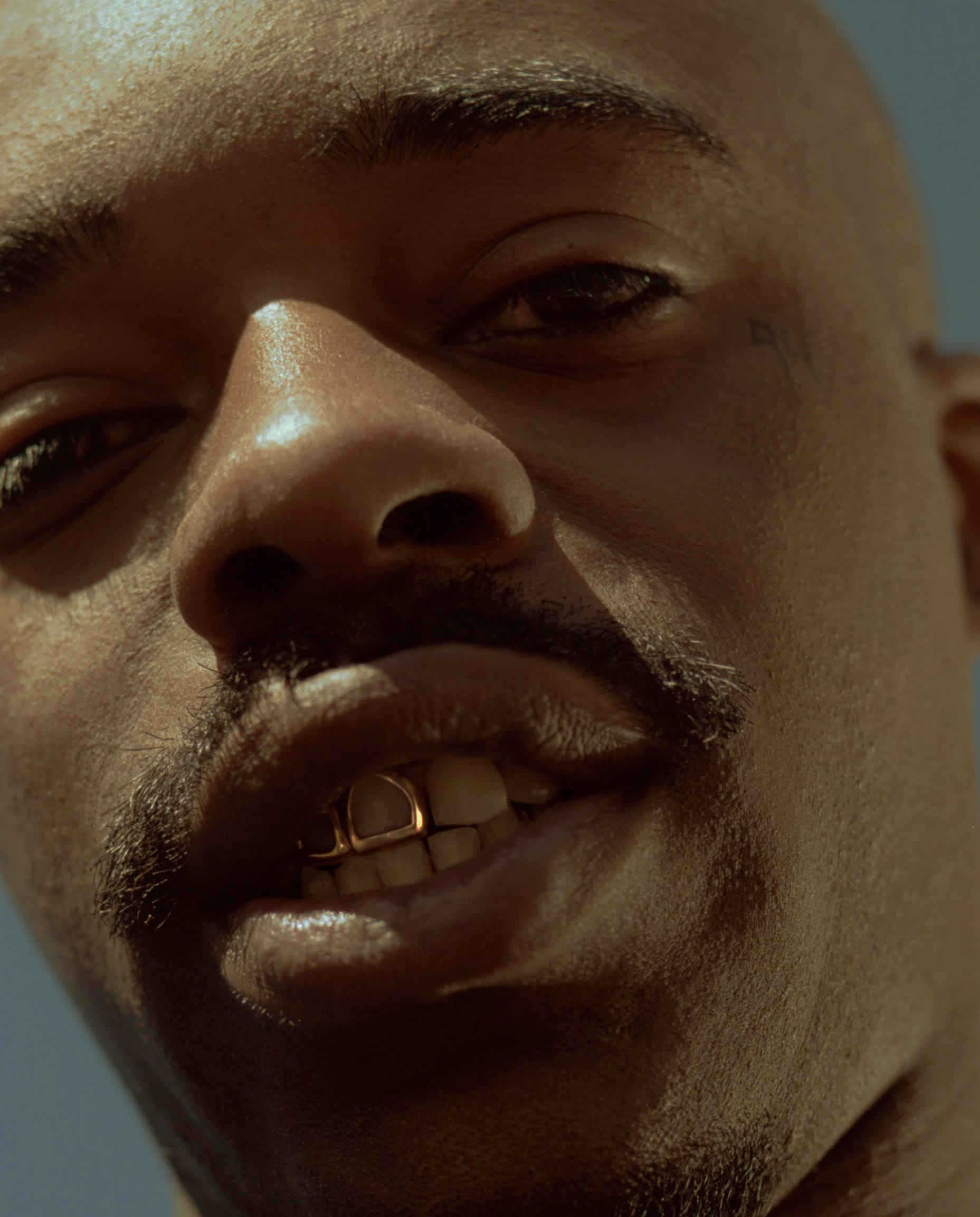 Close-up of a person with a mustache, wearing gold dental jewelry. The background is a clear blue sky.