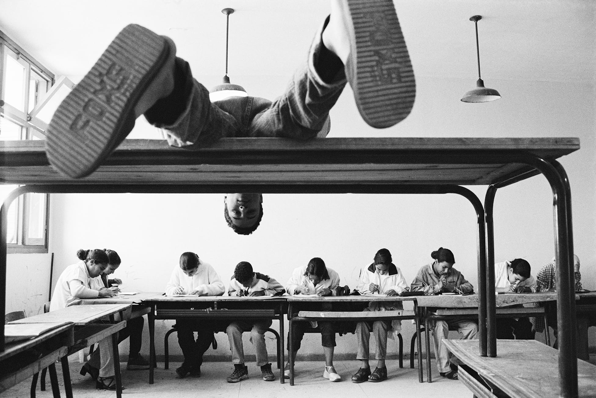 A person lies on top of a table, hanging upside down, while others sit below, focused on writing at their desks in a classroom.
