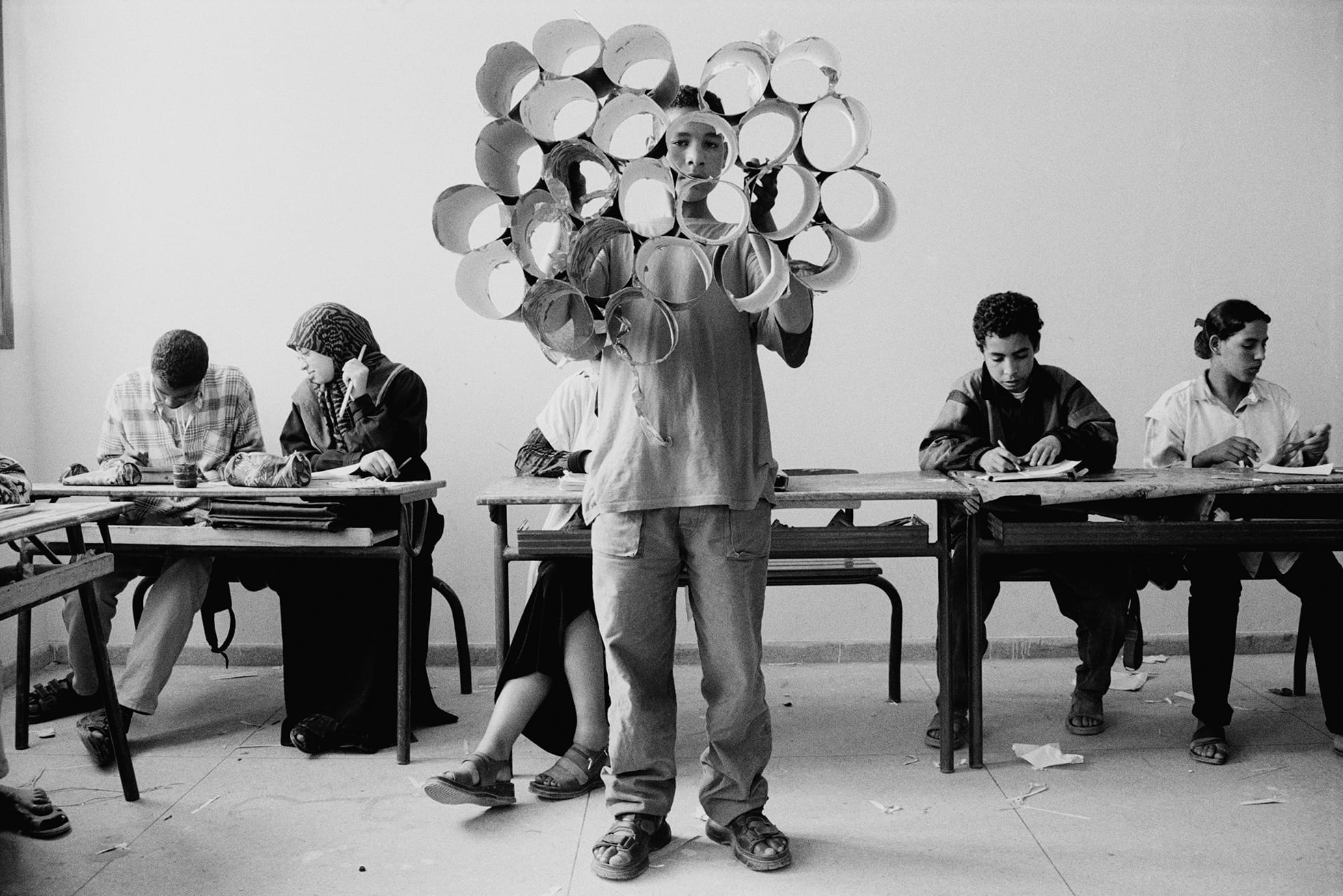 A boy stands holding a circular art piece made of paper rolls in a classroom, while other students sit at desks working on projects.