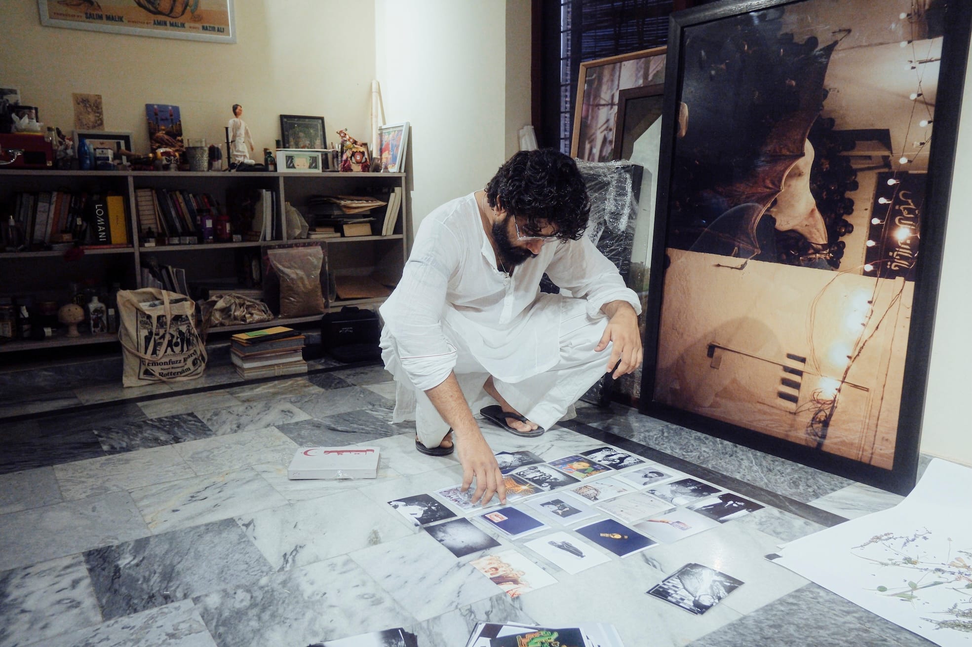 A man in a white outfit squats on a marble floor, organizing photos. Shelves filled with books and decorations are in the background.