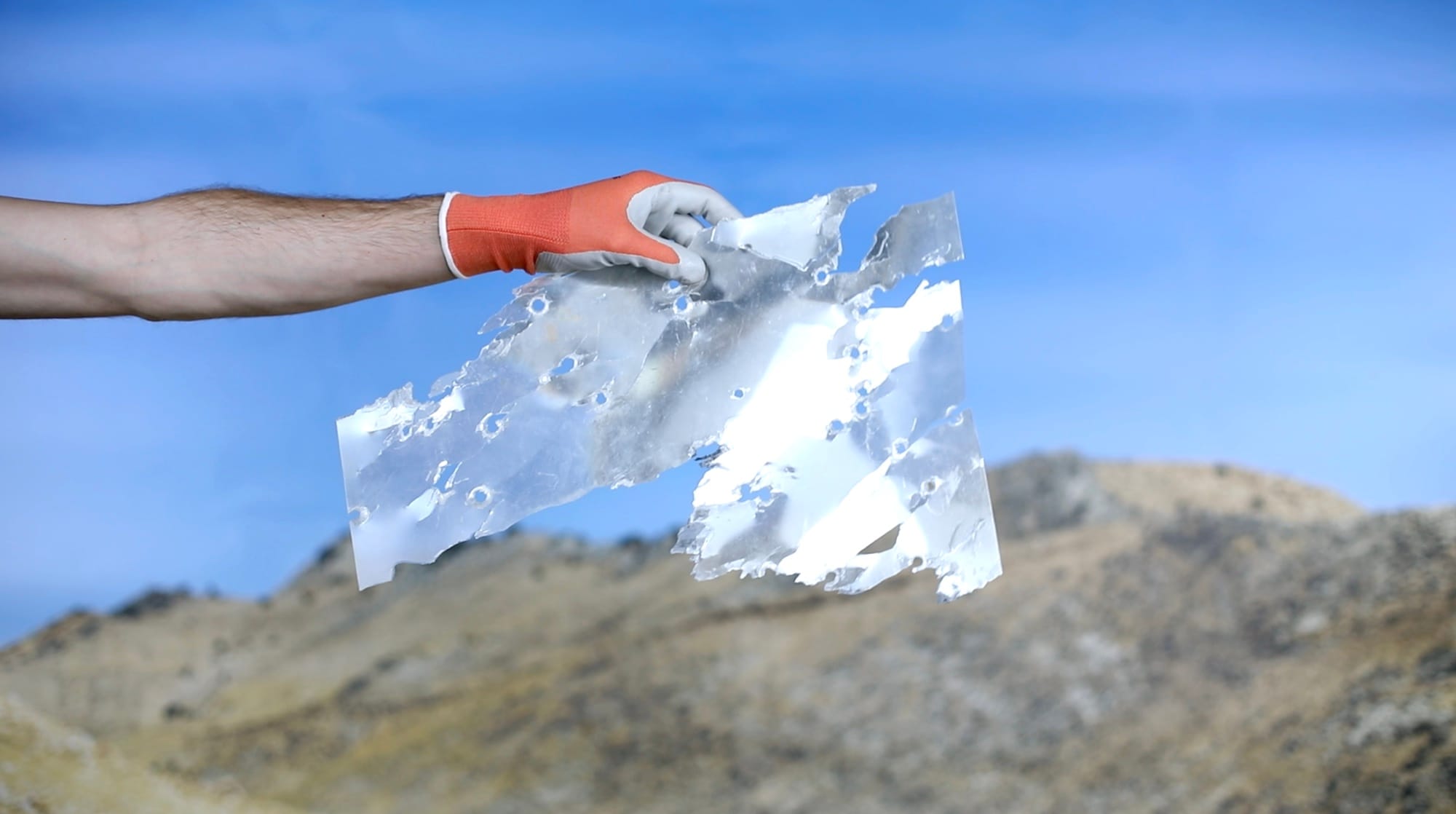 Video still showing a white person's arm entering the frame from the left with an orange glove, holding a piece of gun-shot debris. © Jaclyn Wright