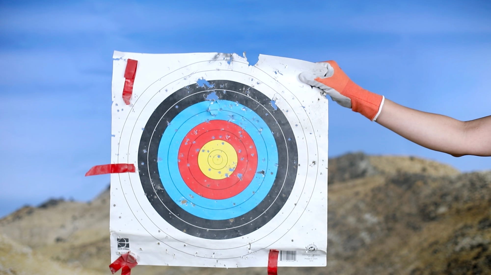 Video still showing a white person's arm entering the frame from the right with an orange glove, holding a piece of gun-shot target paper. © Jaclyn Wright