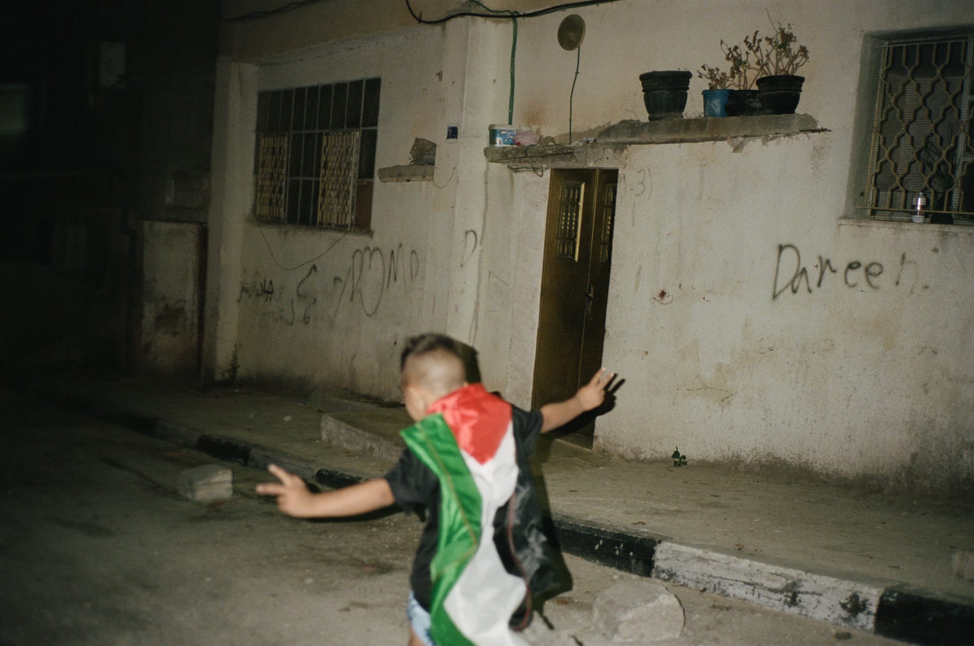 Child runs in dimly lit alley, wearing a flag as a cape. Graffiti covers the wall of a worn building with potted plants above the door.