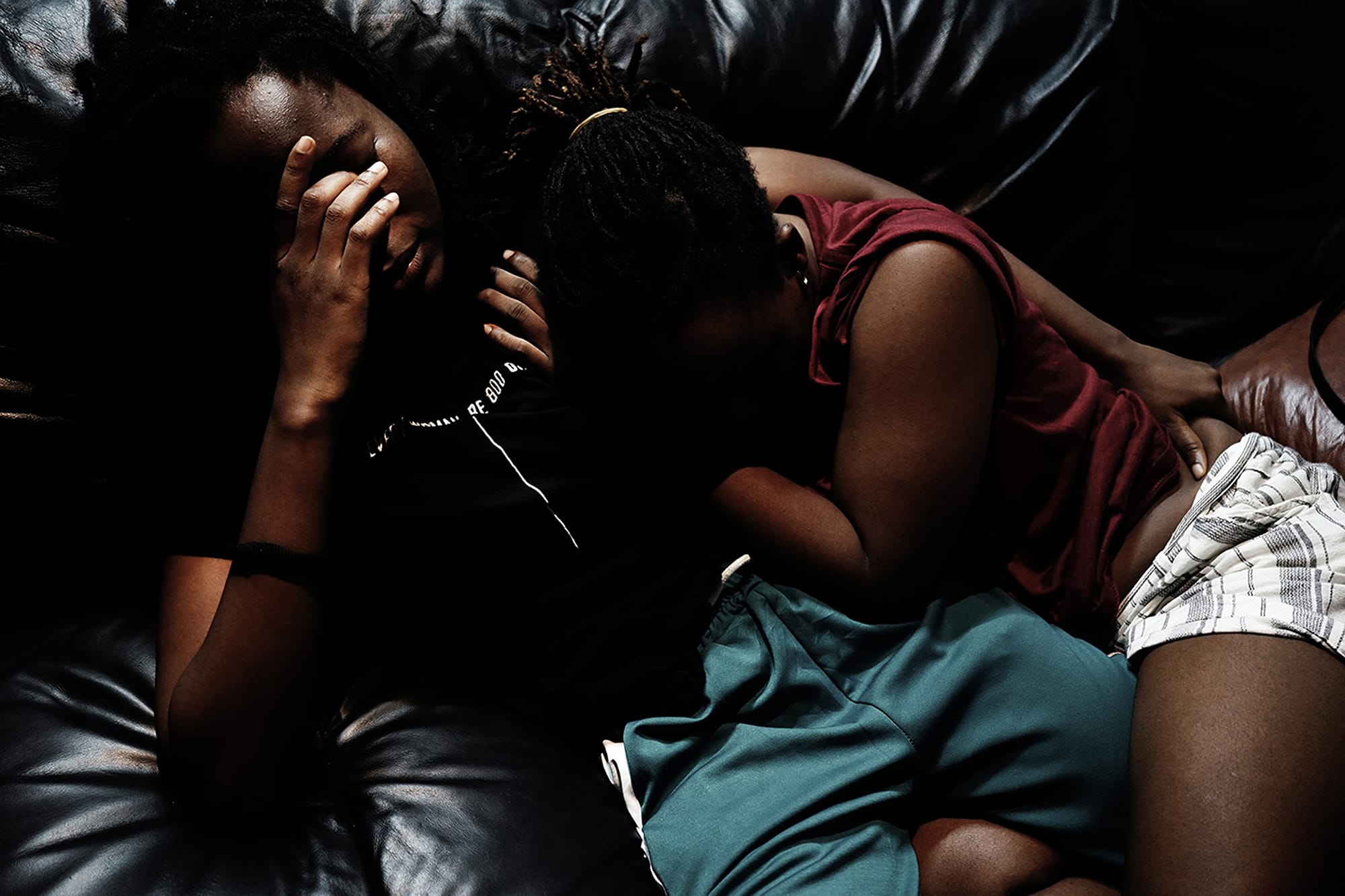 Two people sitting closely on a dark leather couch, one covering their face with their hand, the other leaning against them.