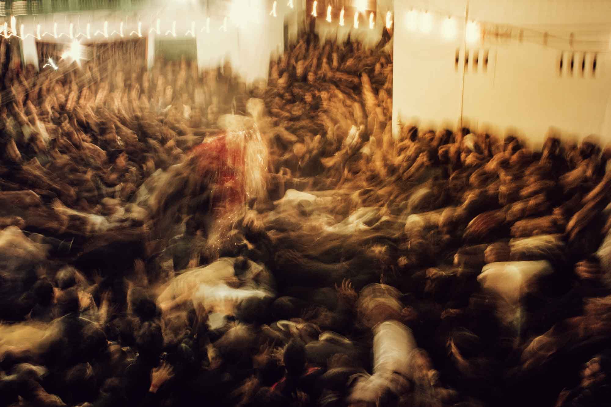 Blurred image of a crowded gathering with people raising their hands in a dimly lit environment.