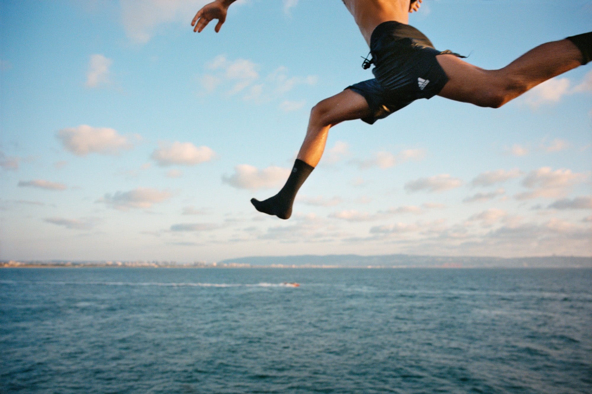 Person in mid-air leap over the ocean, wearing black shorts and socks. Blue sky with clouds in the background.