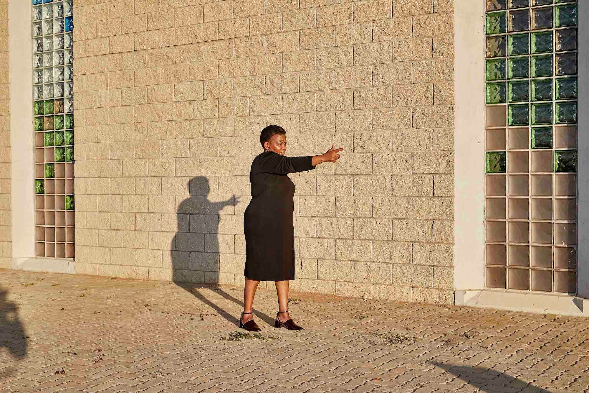 Photograph of a Black woman in front of a stone wall, pointing her finger as if it were a gun © Thero Makepe